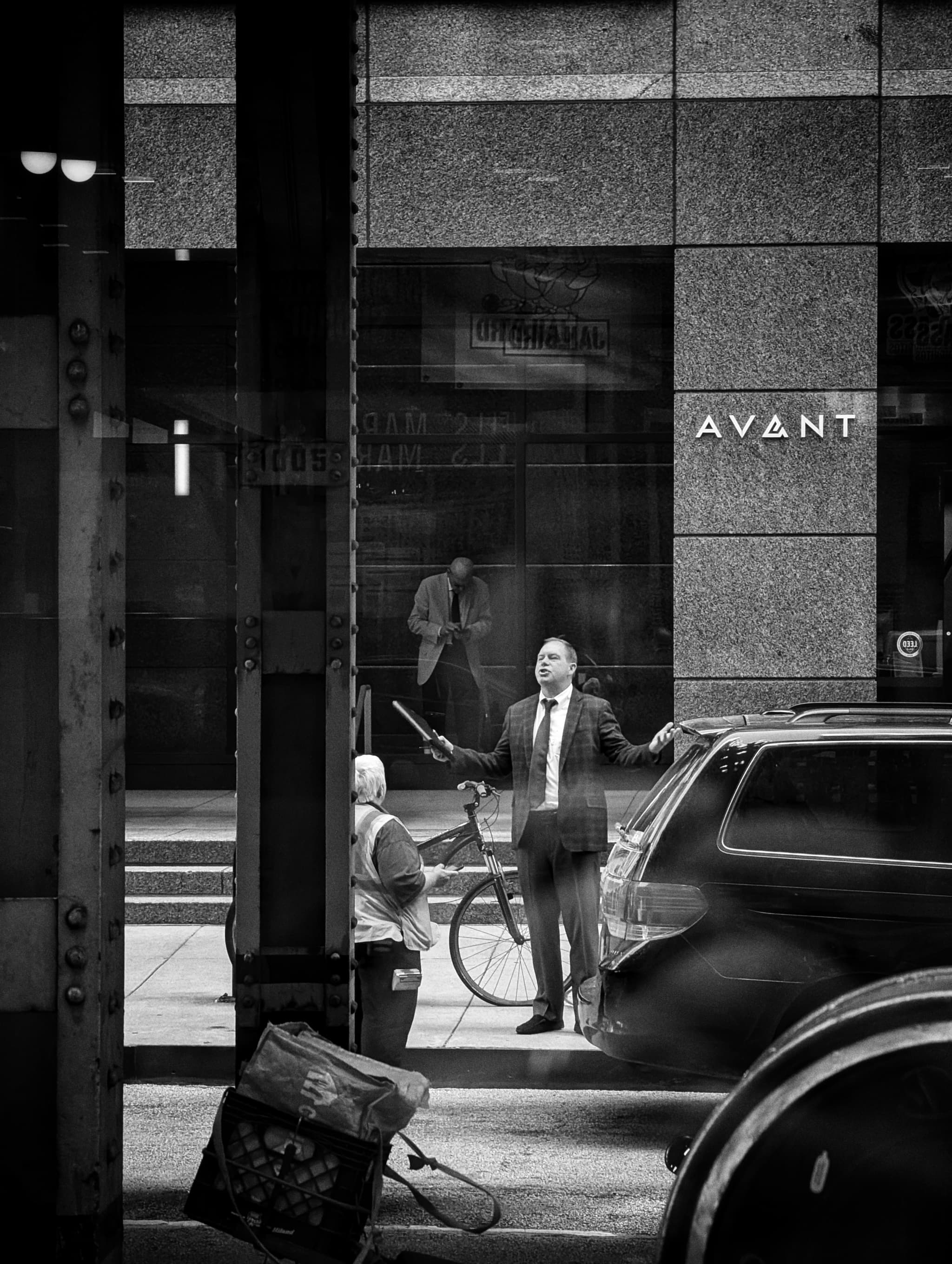 A black and white street scene capturing a man in a suit exiting a vehicle, with another person standing by the car door, and a reflection of a pedestrian visible in the background