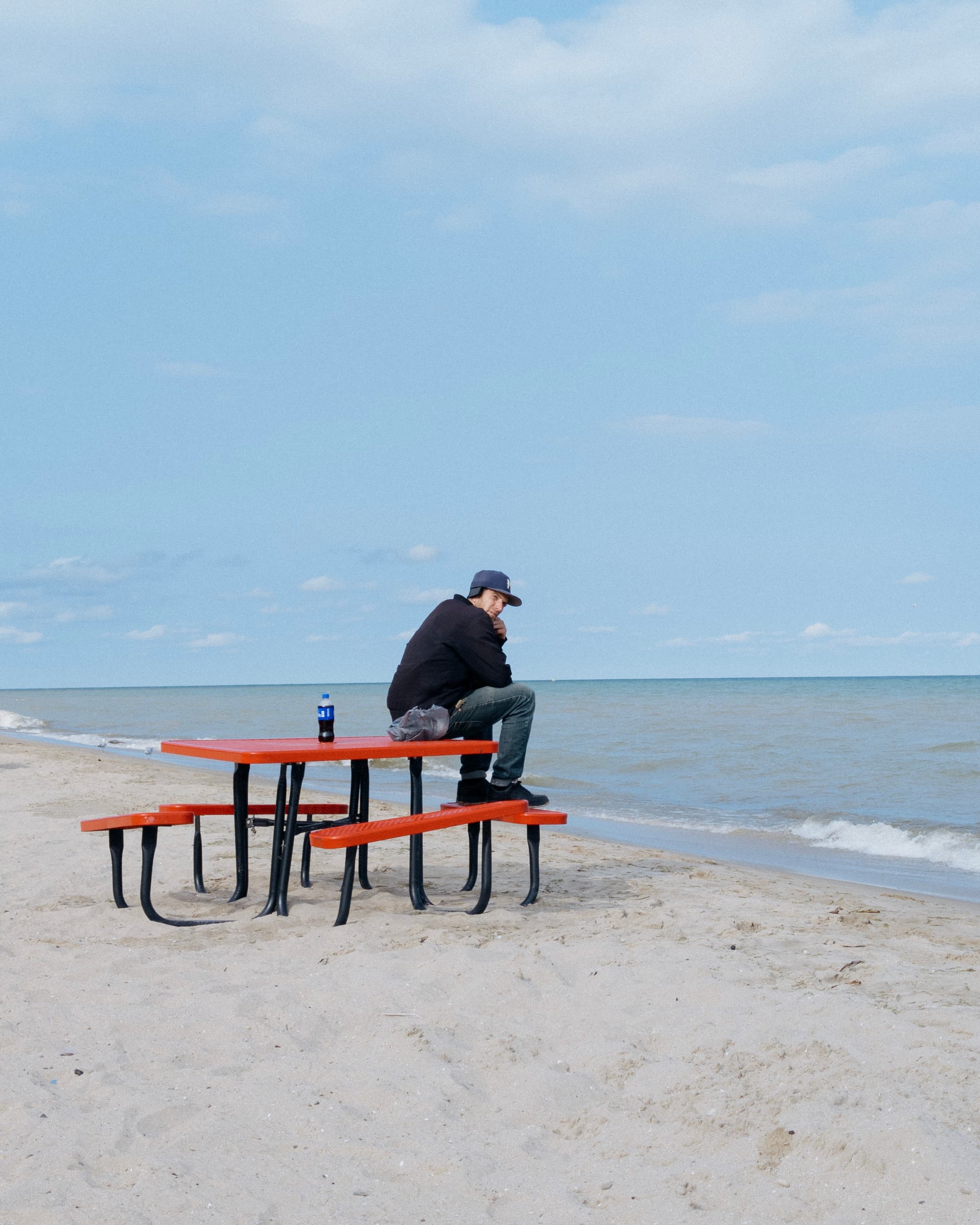 A person sits on a picnic table by the beach, gazing out at the sea with a beverage can beside them