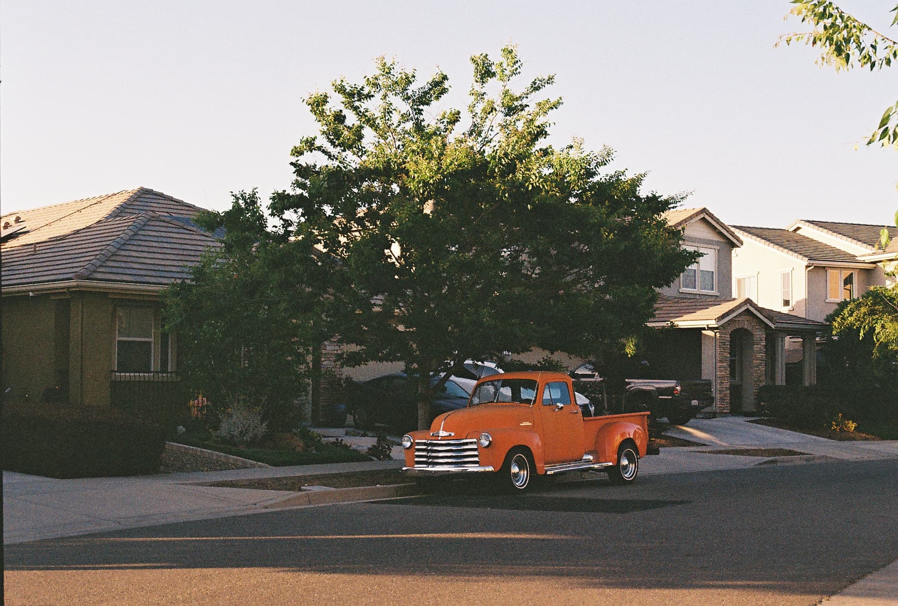 A vintage orange truck parked on a suburban street with houses and trees in the background