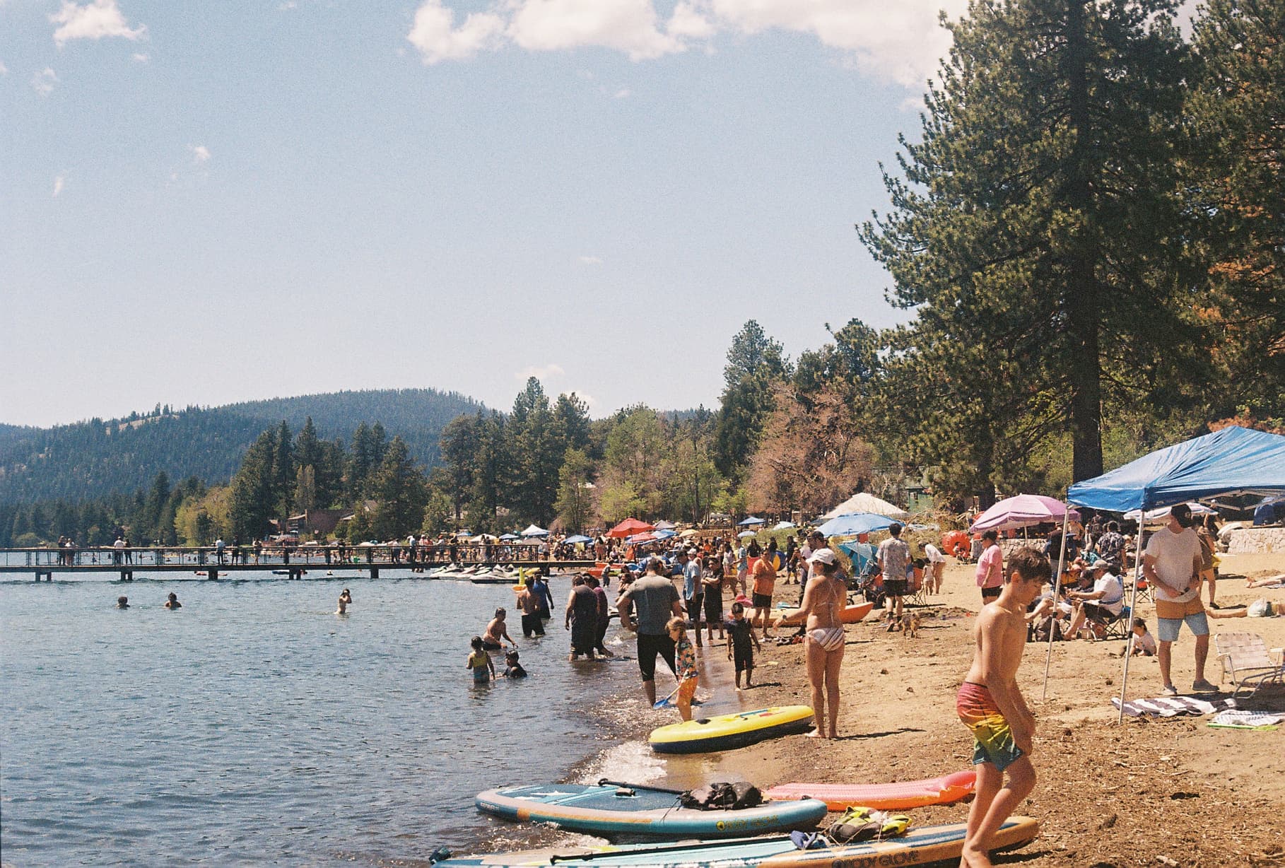 A bustling lakeside scene with people swimming, sunbathing, and engaging in various activities on a sunny day, with trees and hills in the background