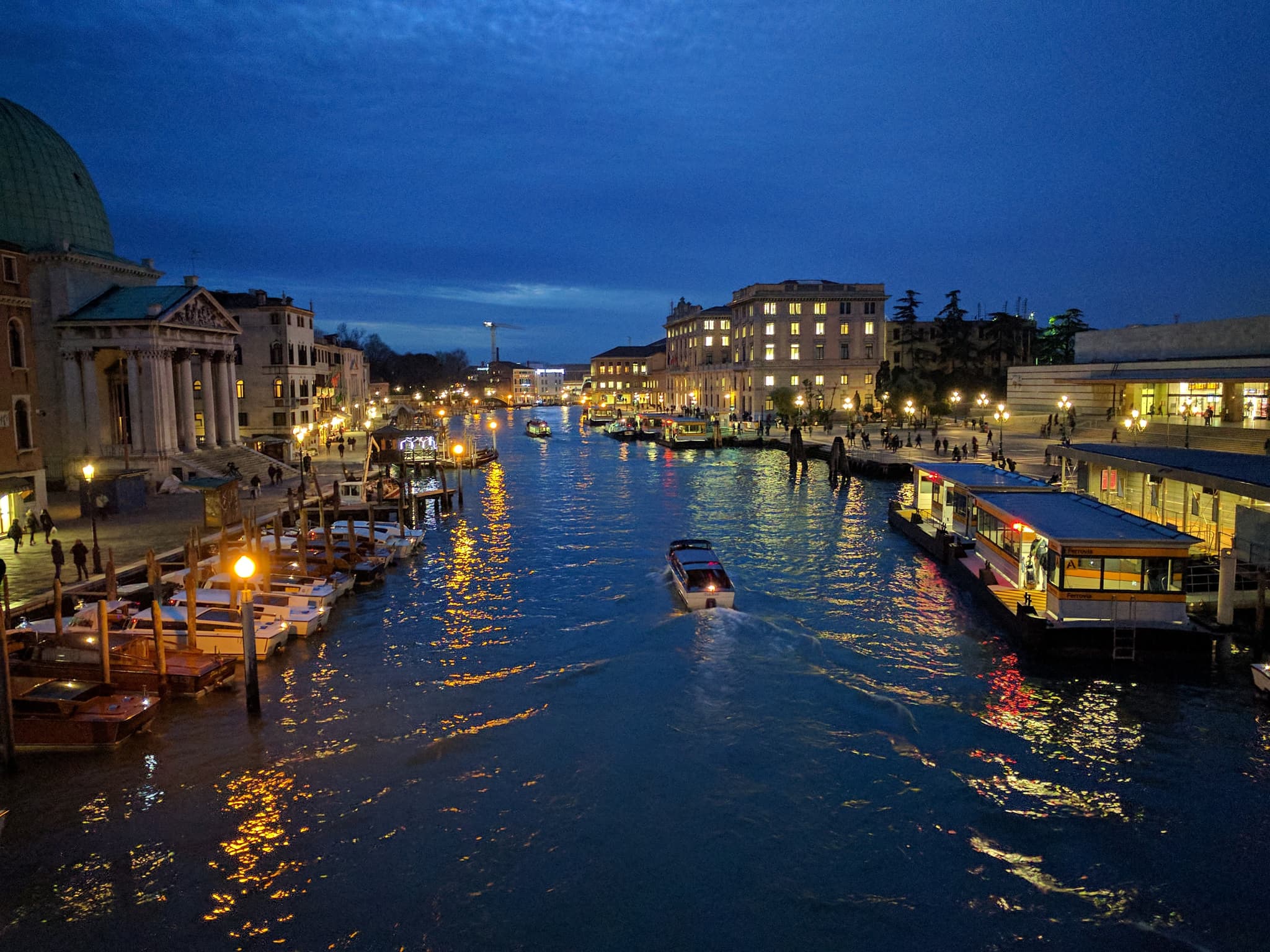 A twilight scene over a bustling canal with boats, adjacent buildings illuminated by warm lights, and a clear blue sky transitioning to night