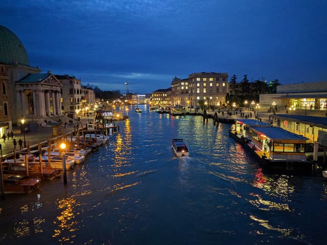 A twilight scene over a bustling canal with boats, adjacent buildings illuminated by warm lights, and a clear blue sky transitioning to night